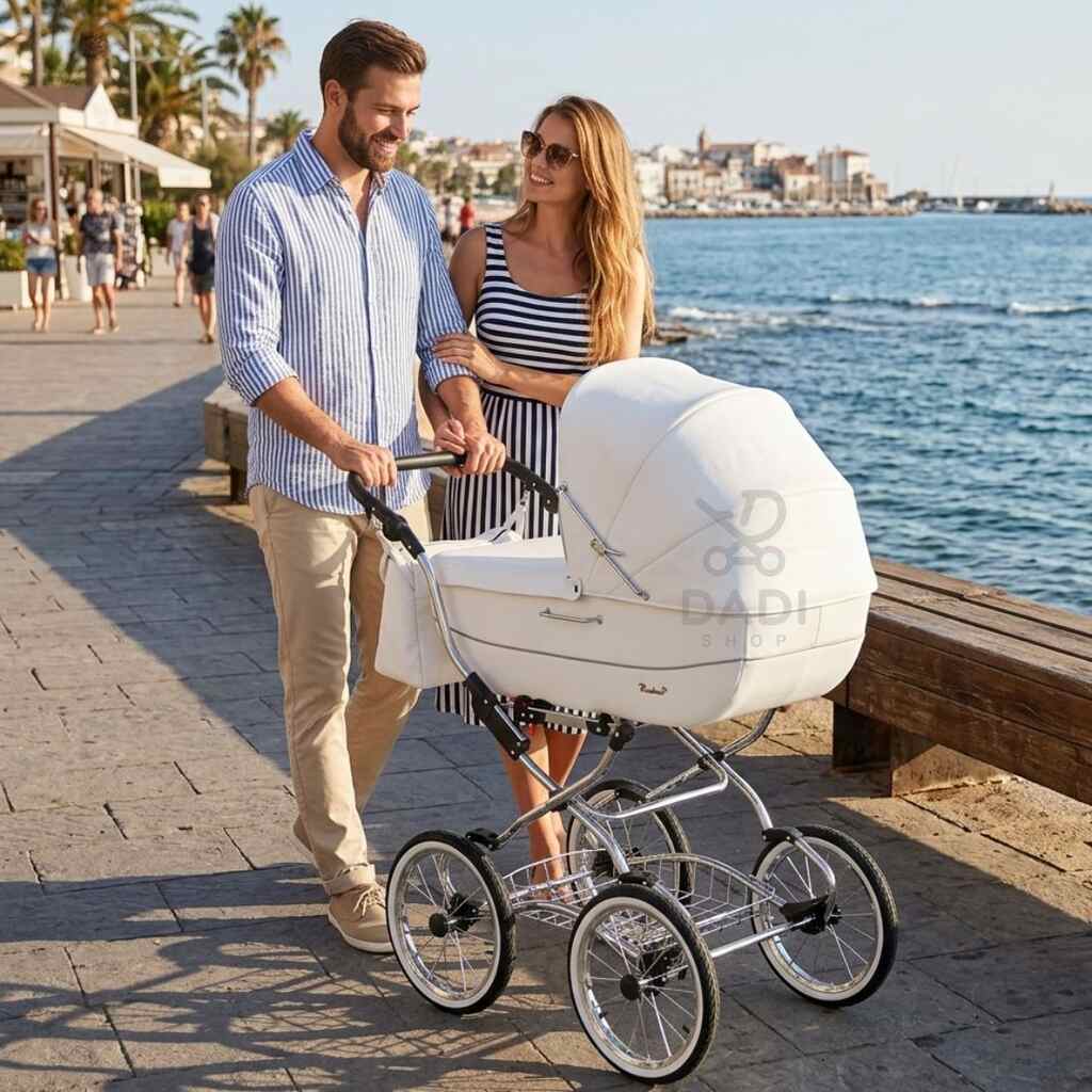 Uśmiechnięta para prowadzi biały wózek Baby Fashion Renee na nadmorskiej promenadzie w słoneczny dzień.