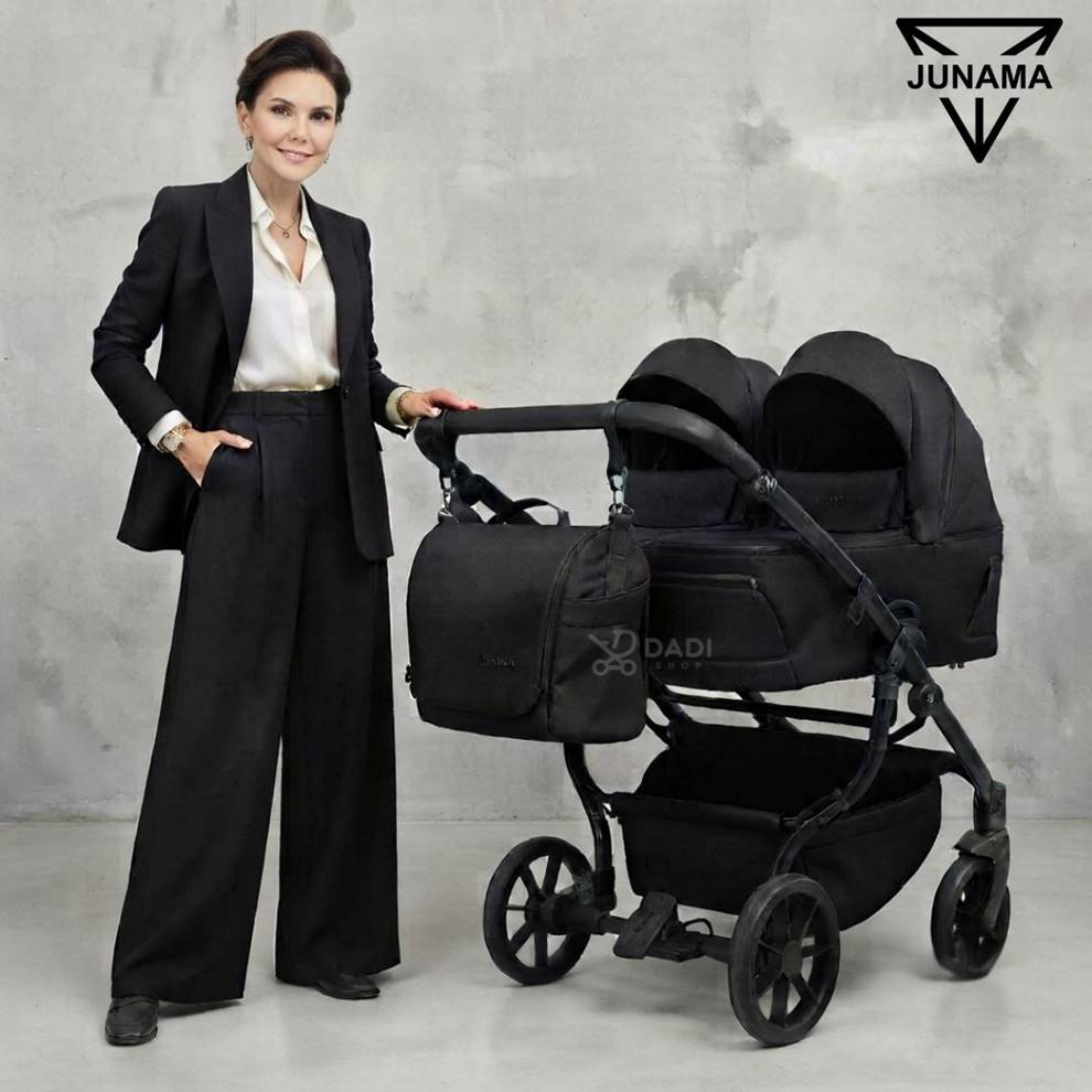 Modelka obok wózka dla bliźniąt Junama Duo V4 w kolorze czarnym. Zestaw 2w1 z torbą pielęgnacyjną w studio na szarym tle.