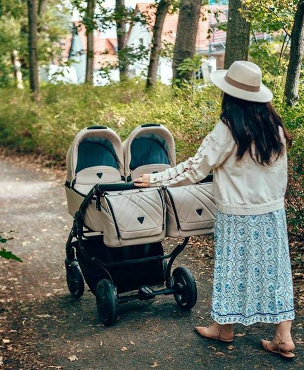 Wózek dla bliźniąt Junama Duo V3 Eco Beige – spacer w parku, kobieta w kapeluszu prowadzi podwójny wózek.