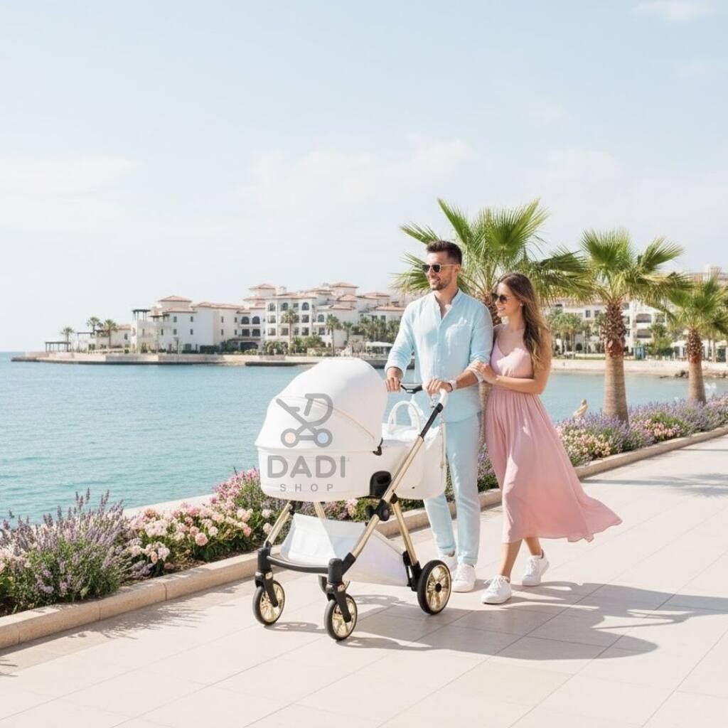 Para na spacerze nad morzem z białym wózkiem Junama Ellegante Art Deco. Słoneczna sceneria z palmami i luksusową architekturą.