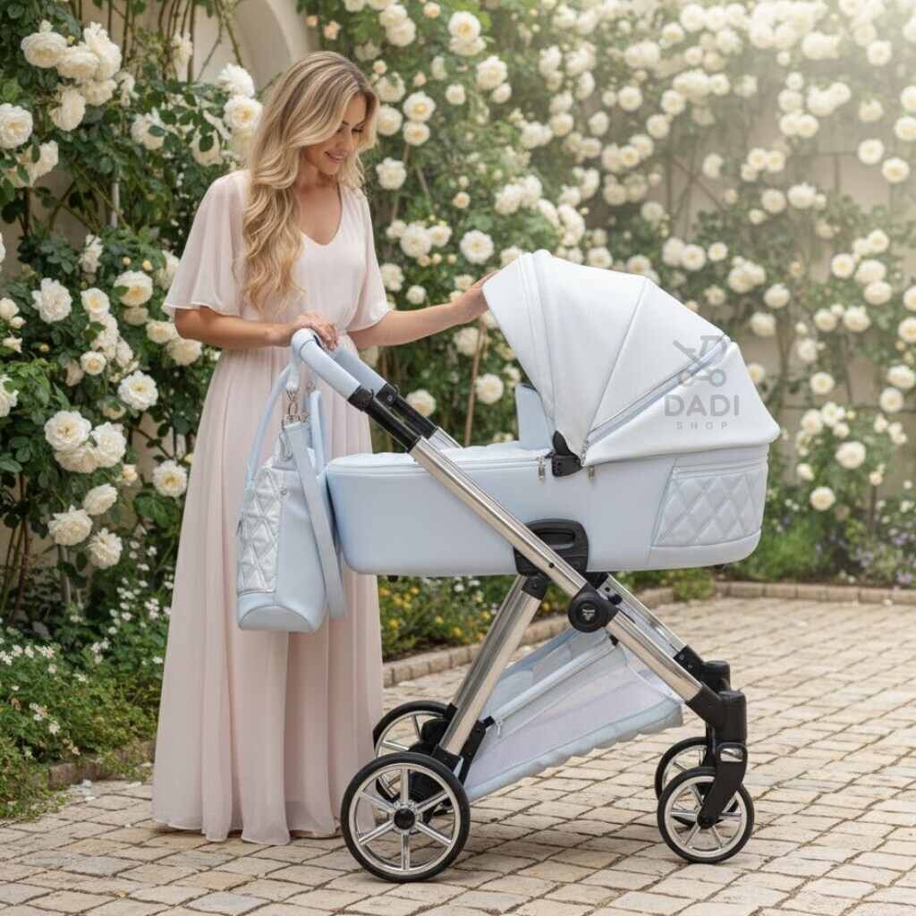 Elegancka kobieta w jasnej sukni w ogrodzie białych róż, z jasnoniebieskim wózkiem-gondolą Junama Ellegante Lame 03. Wózek ma błyszczącą, pikowaną tapicerkę i torbę