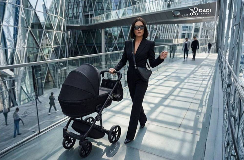Confident woman walking while pushing a sleek black Junama Elegante pram. Urban glass architecture in the background.