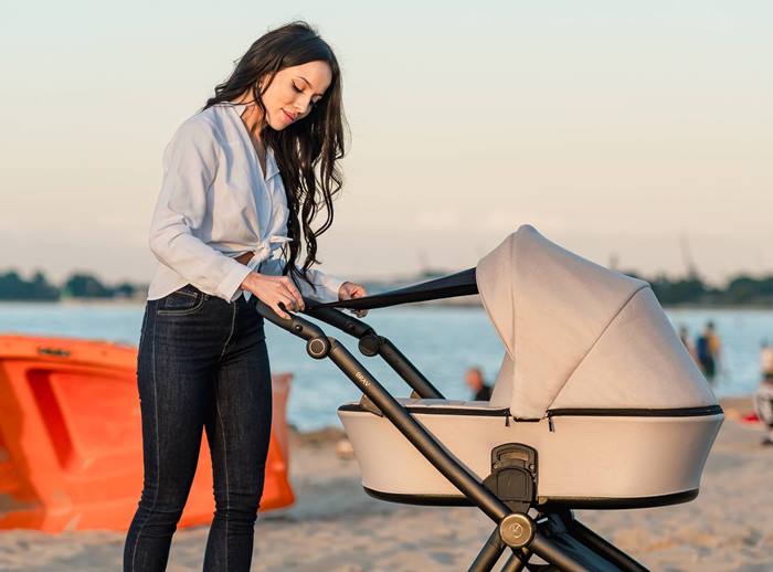Gondola wózka BRAV z budką przeciwsłoneczną – mama montuje blendę na stelażu podczas spaceru nad plażą.
