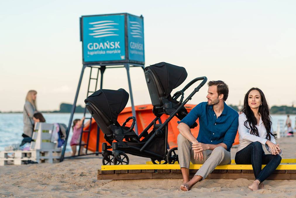 Kobieta i mężczyzna siedzący na plaży w Gdańsku koło wózka podwójnego Kunert Ivento Duo z dwoma siedziskami spacerowymi.