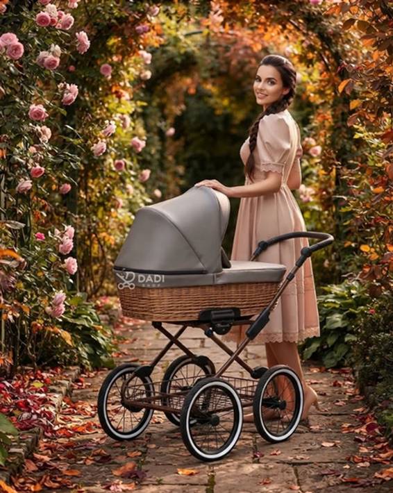 Wózek dziecięcy 2w1 Kunert Romantic Classic w stylu retro z wiklinową gondolą i szarą budką, stojący w alei różanej z kwitnącymi krzewami, obok którego stoi uśmiechnięta kobieta