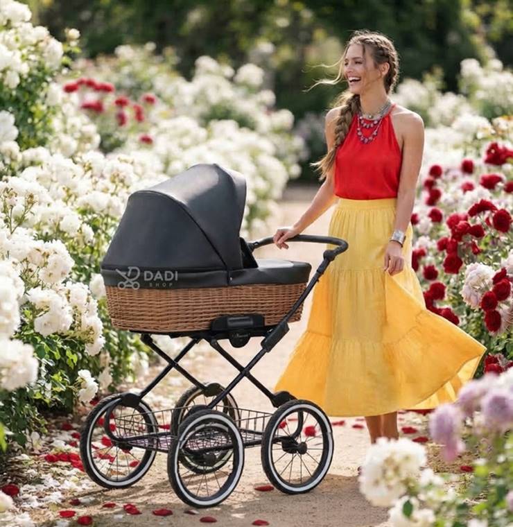 Uśmiechnięta kobieta w czerwonym topie i żółtej spódnicy pcha wózek Kunert Romantic Classic z wiklinową gondolą w alei pełnej kwitnących białych i czerwonych róż.