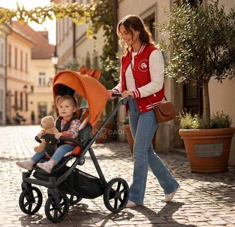 Mama spaceruje z córką w wózku spacerowym Natoni Joy w kolorze Fox (pomarańczowym) po słonecznej, wybrukowanej uliczce.