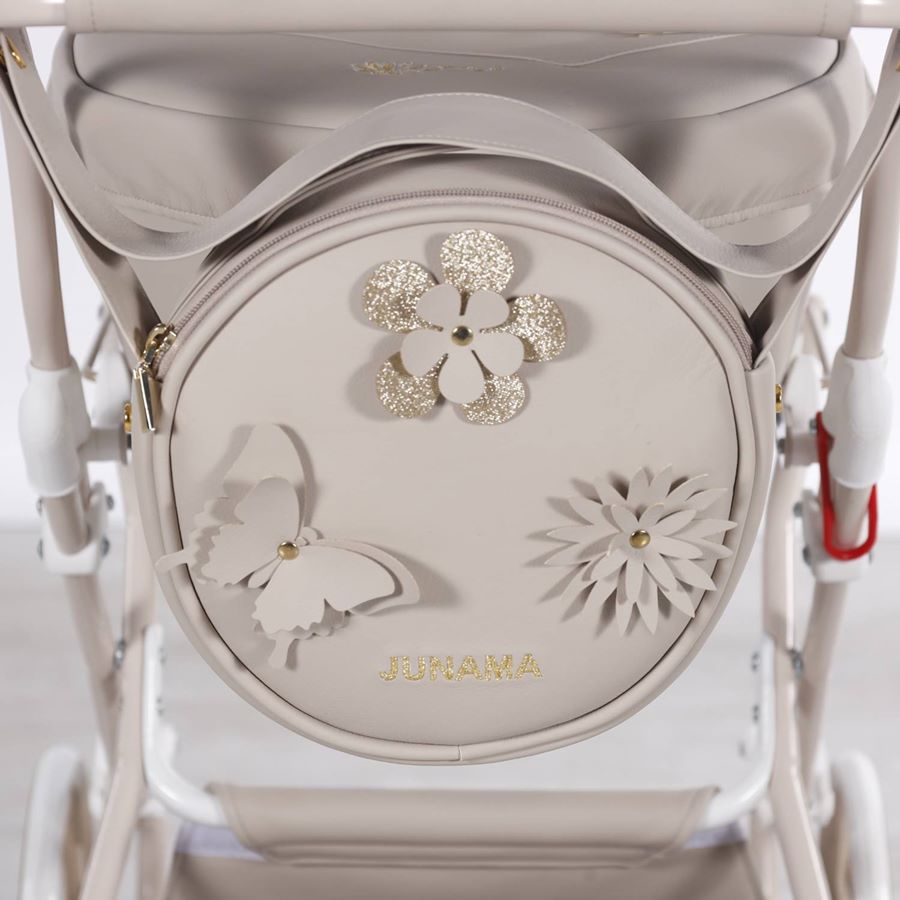 Zbliżenie na okrągłą, beżową torbę z błyszczącymi kwiatami, motylkiem i logo Junama, zawieszoną na rączce wózka dla lalek Fleur Mini.