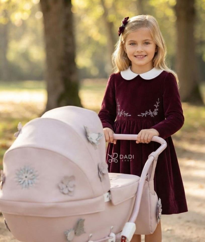 A little girl in a burgundy dress strolls with a pink Junama Mini Fleur doll pram in an autumn park.