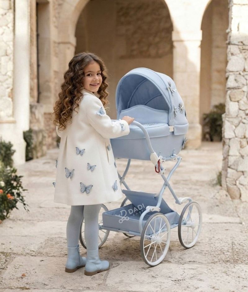 Dziewczynka w płaszczyku z motylami prowadzi błękitny wózek Junama Fleur Mini Blue na zabytkowym, kamiennym patio.