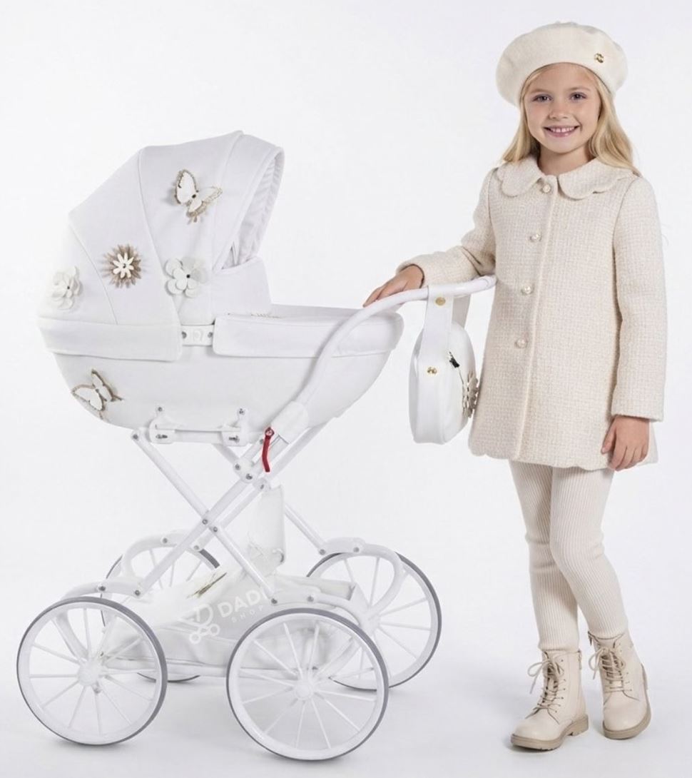 Biały wózek dla lalek Junama Fleur Mini 04 z ozdobnymi kwiatami i motylami, na retro stelażu. Obok stoi uśmiechnięta dziewczynka w berecie.