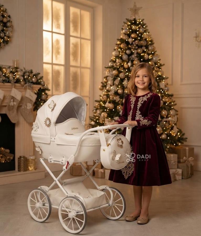 Odświętnie ubrana dziewczynka pozuje przy oświetlonej choince obok luksusowego, białego wózka dla lalek Junama Mini Fleur.