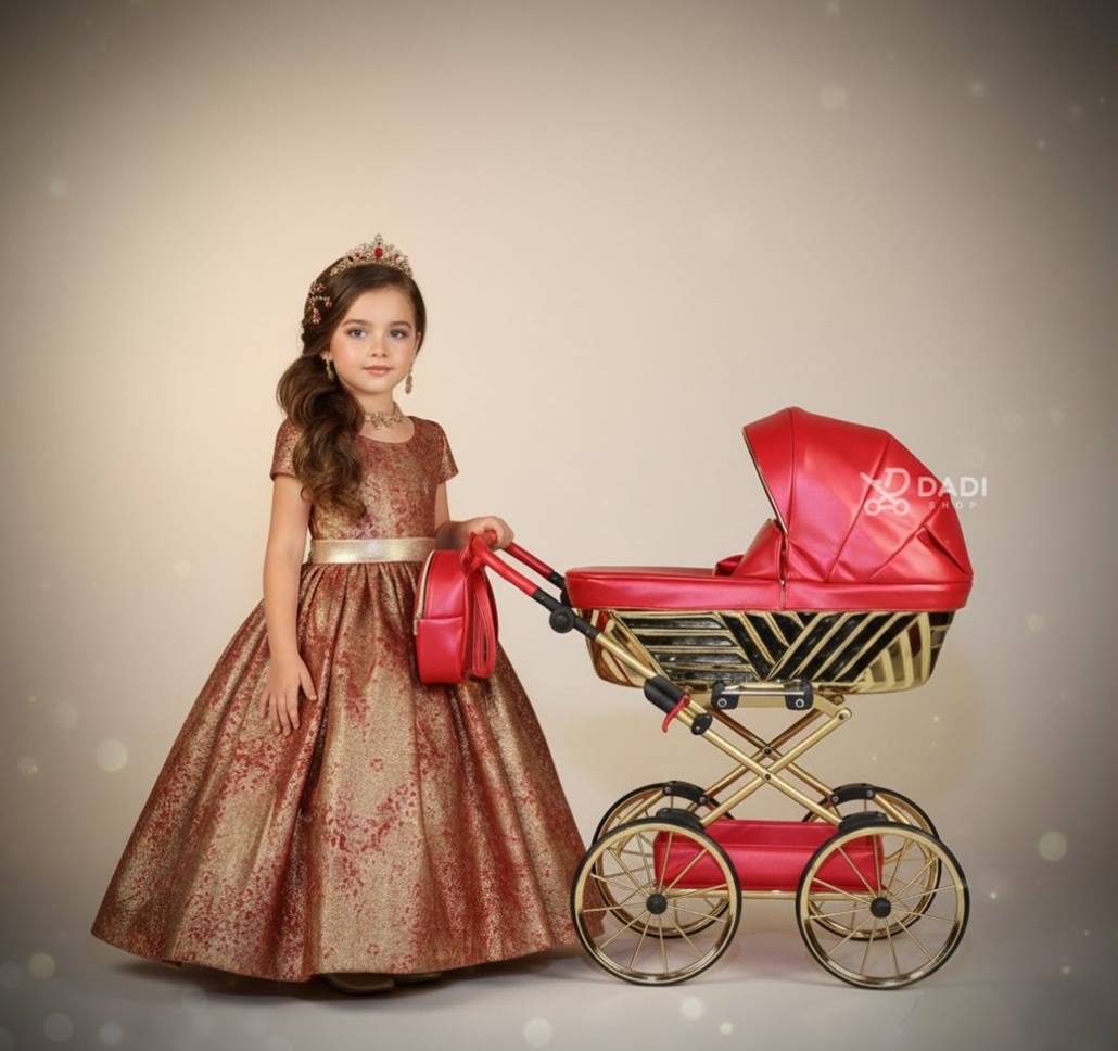 Luksusowy wózek dla lalek Royal w złoto-czerwonym kolorze, prezentowany przez 5-letnią dziewczynkę w zimowej sukni haute couture.